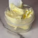 Close-up of creamy homemade butter in a small glass jar with a spatula, showing smooth, freshly whipped texture.