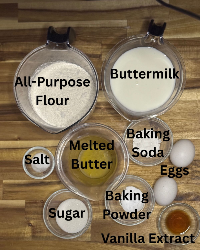 Ingredients for easy buttermilk pancakes from scratch including all-purpose flour, buttermilk, melted butter, eggs, sugar, baking powder, baking soda, salt, and vanilla extract arranged in glass bowls on a wooden surface.