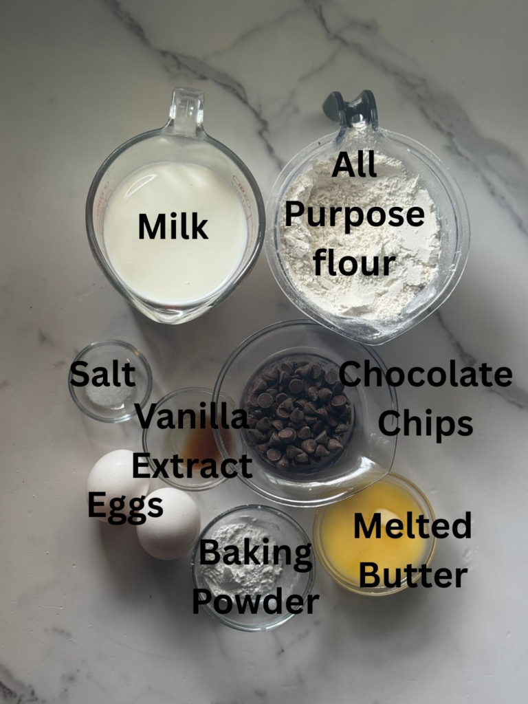 Ingredients for homemade chocolate chip pancakes including milk, all-purpose flour, eggs, melted butter, baking powder, vanilla extract, salt, and chocolate chips arranged on a marble countertop.
