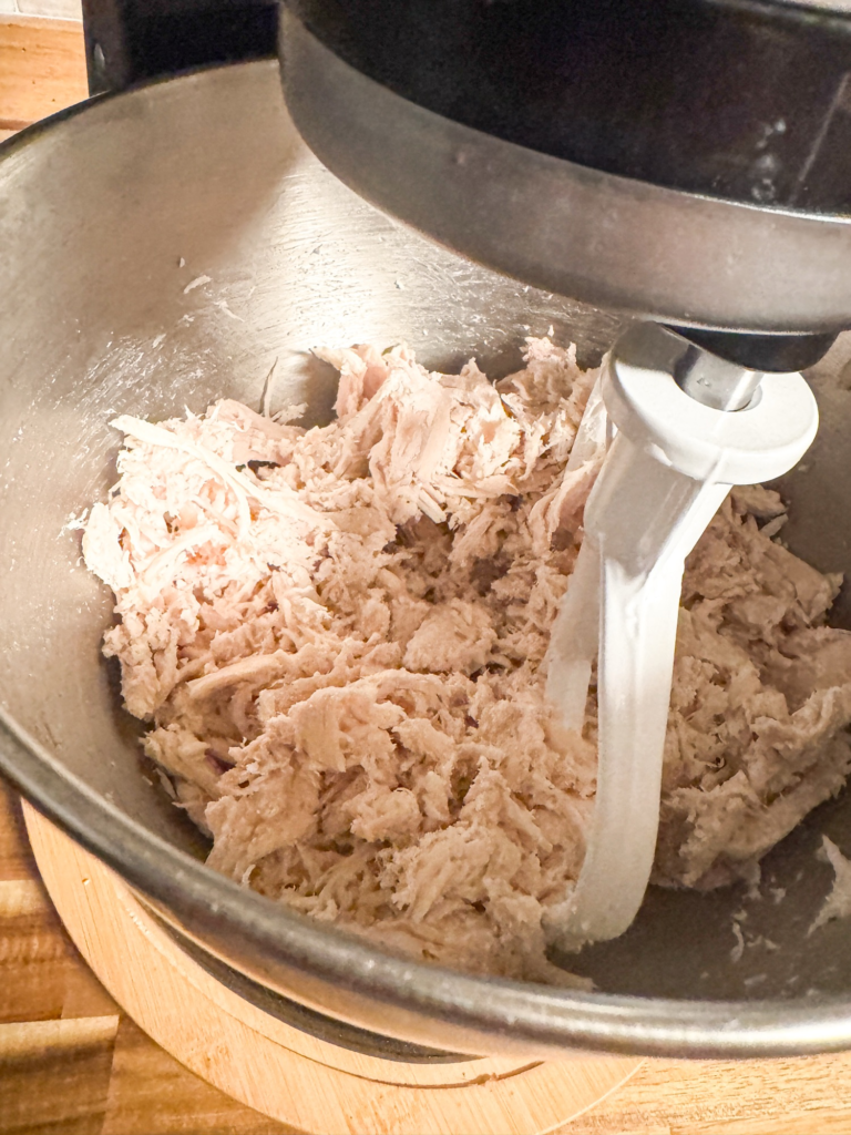 Cooked chicken being shredded in a stand mixer with paddle attachment for chicken salad