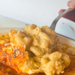 This close-up shows a spoonful of creamy baked mac and cheese being lifted from a casserole dish. The pasta is coated in a rich, smooth cheese sauce while the top is baked to a golden, slightly crispy finish. The combination of melty cheese and a lightly browned crust gives this dish the perfect balance of creamy and crunchy textures.