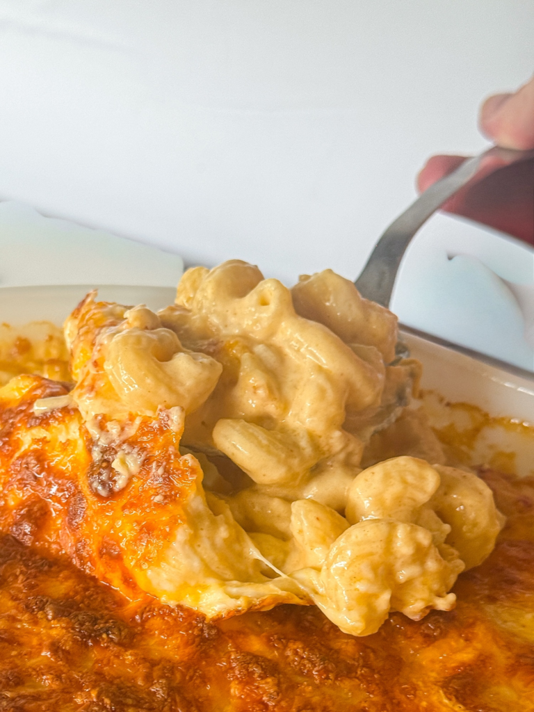This close-up shows a spoonful of creamy baked mac and cheese being lifted from a casserole dish. The pasta is coated in a rich, smooth cheese sauce while the top is baked to a golden, slightly crispy finish. The combination of melty cheese and a lightly browned crust gives this dish the perfect balance of creamy and crunchy textures.