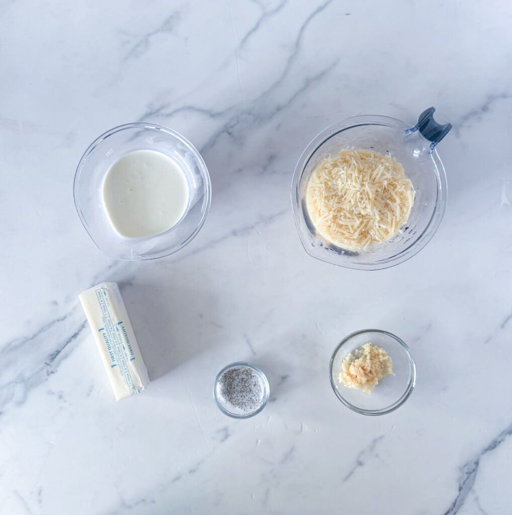 Ingredients for easy homemade Alfredo sauce including butter, heavy cream, parmesan cheese, garlic, salt, and pepper on a marble surface