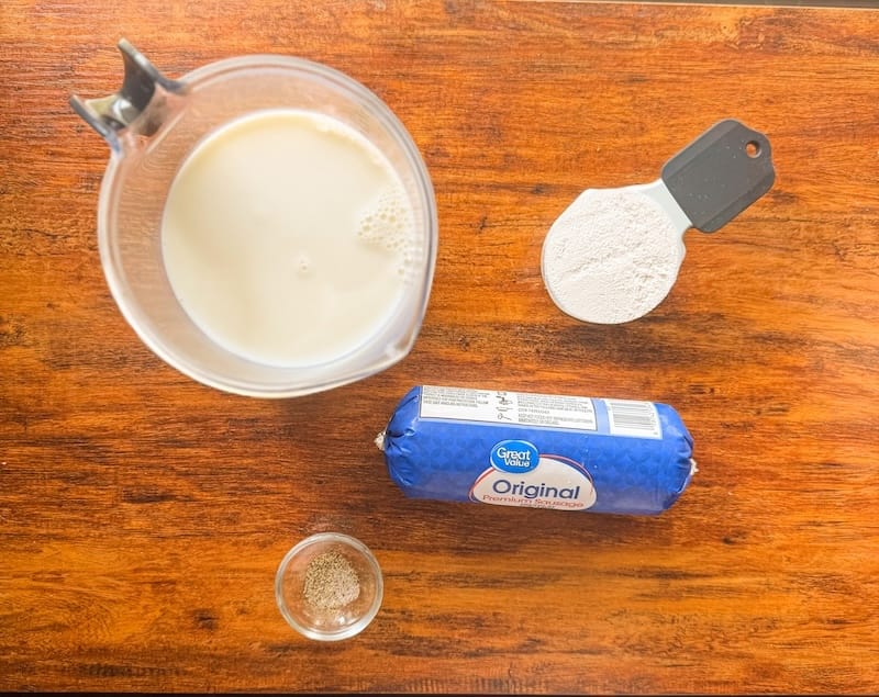 Ingredients for homemade sausage gravy including breakfast sausage, milk, flour, and seasoning on a wooden surface
